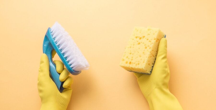 Yellow Background with pair of hands in yellow rubber gloves. One hand is holding a blue scrubbing brush, and the other is holding a yellow rectangular sponge.