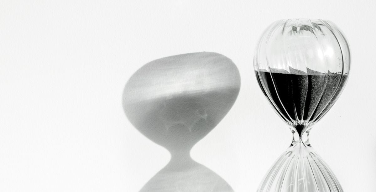 An hourglass with grey sand in the top and a white background with the shadow of the timer on it